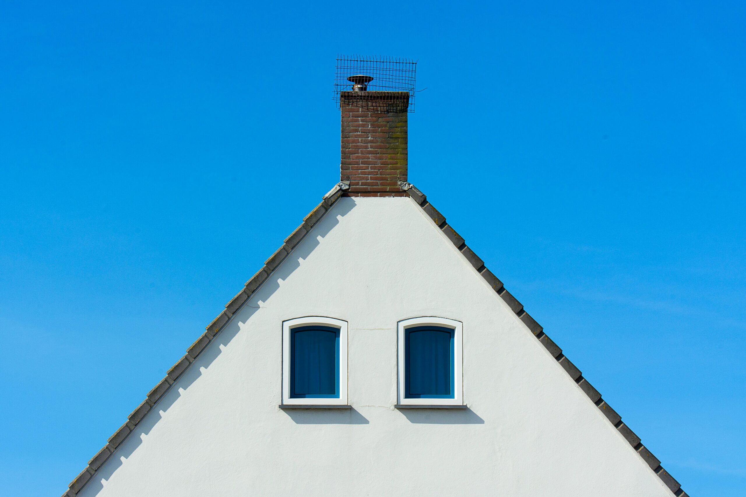 pexels-jan-van-der-wolf-11680885-12426087 A white triangular gable of a house with two arched windows and a brick chimney at the top, covered with a protective metal mesh, set against a bright blue sky.